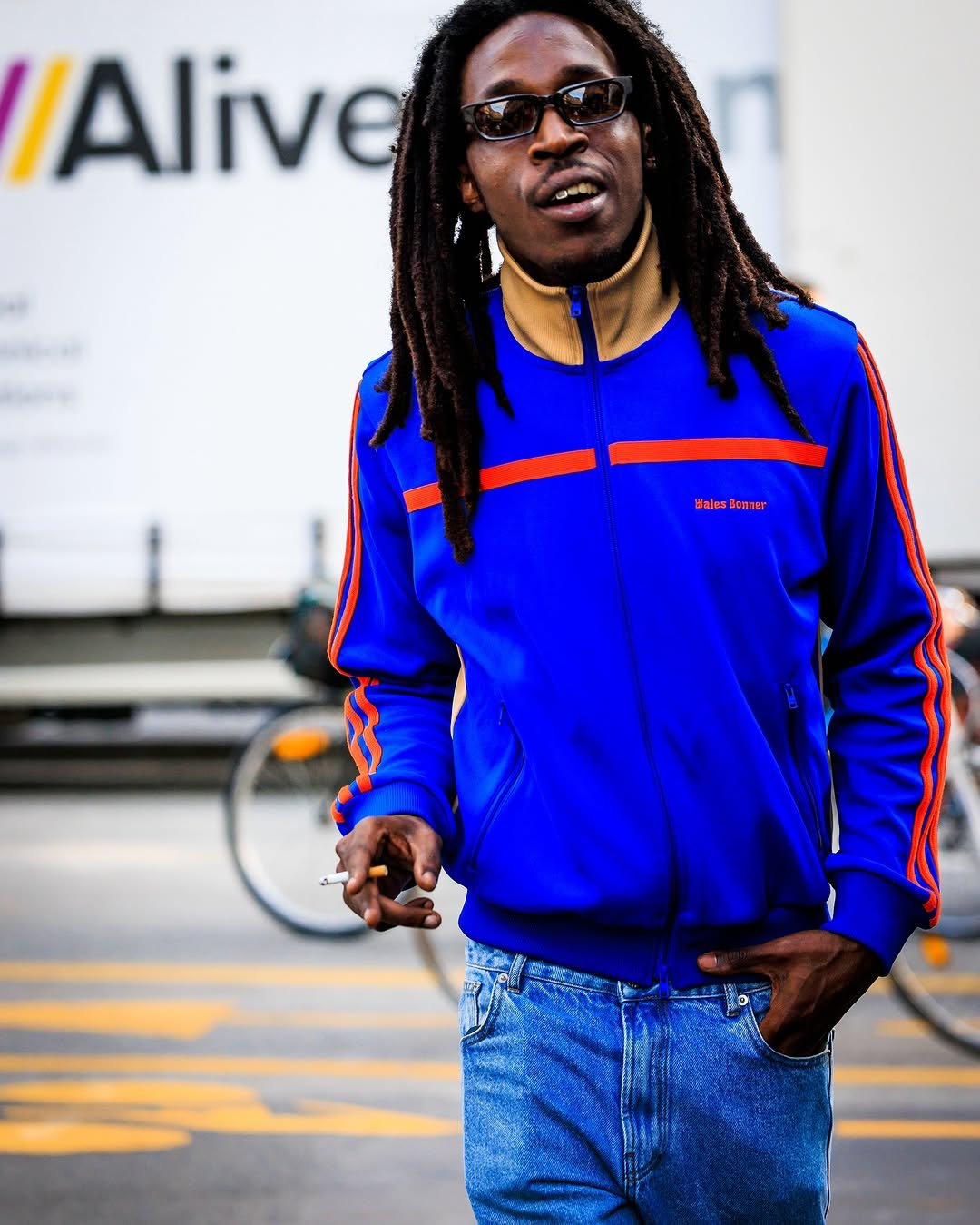 A bright blue track jacket with orange striping is worn over light blue jeans and narrow black sunglasses.