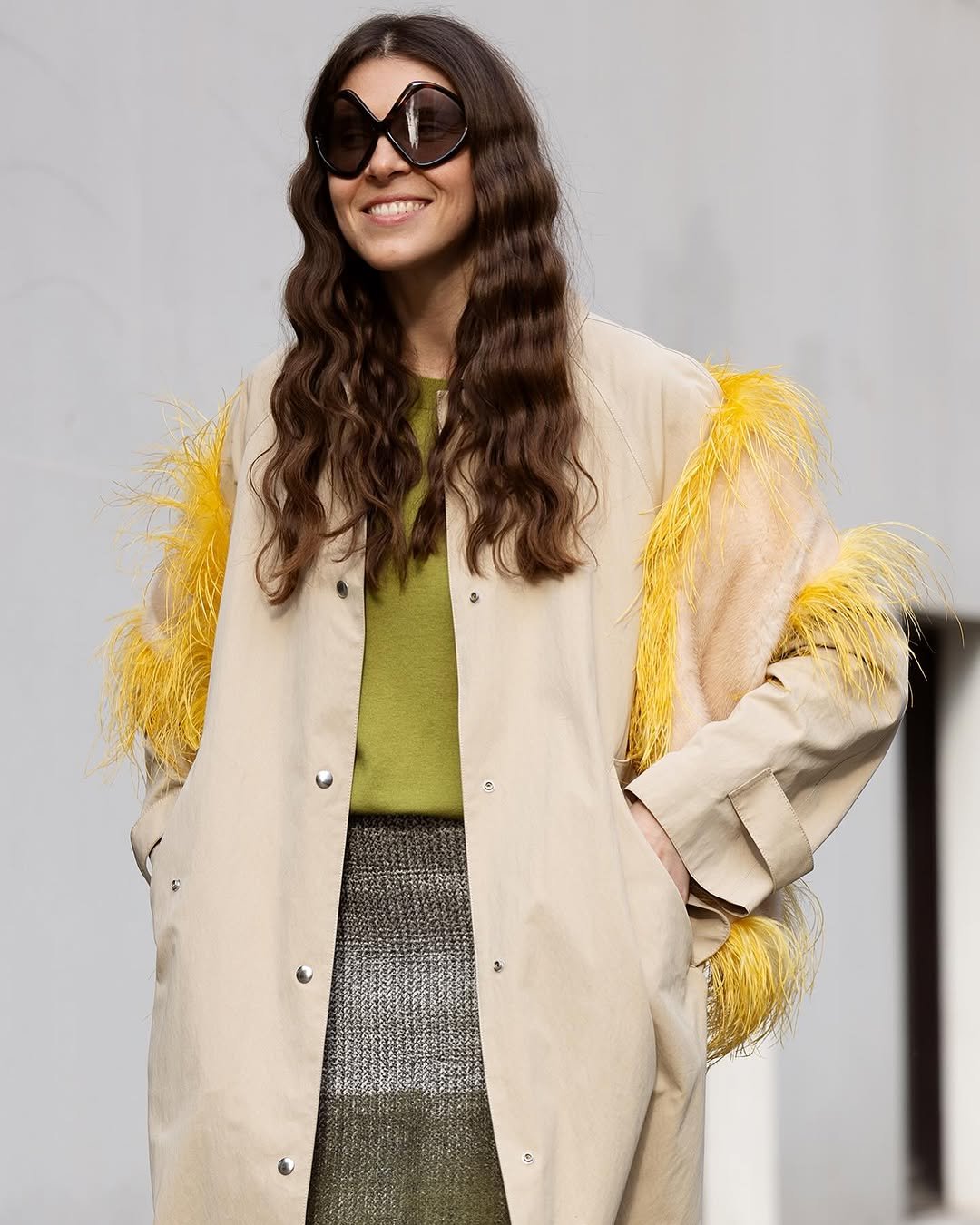 A cream oversized coat with yellow feather-trimmed sleeves is worn over an olive top and textured skirt with oversized sunglasses.