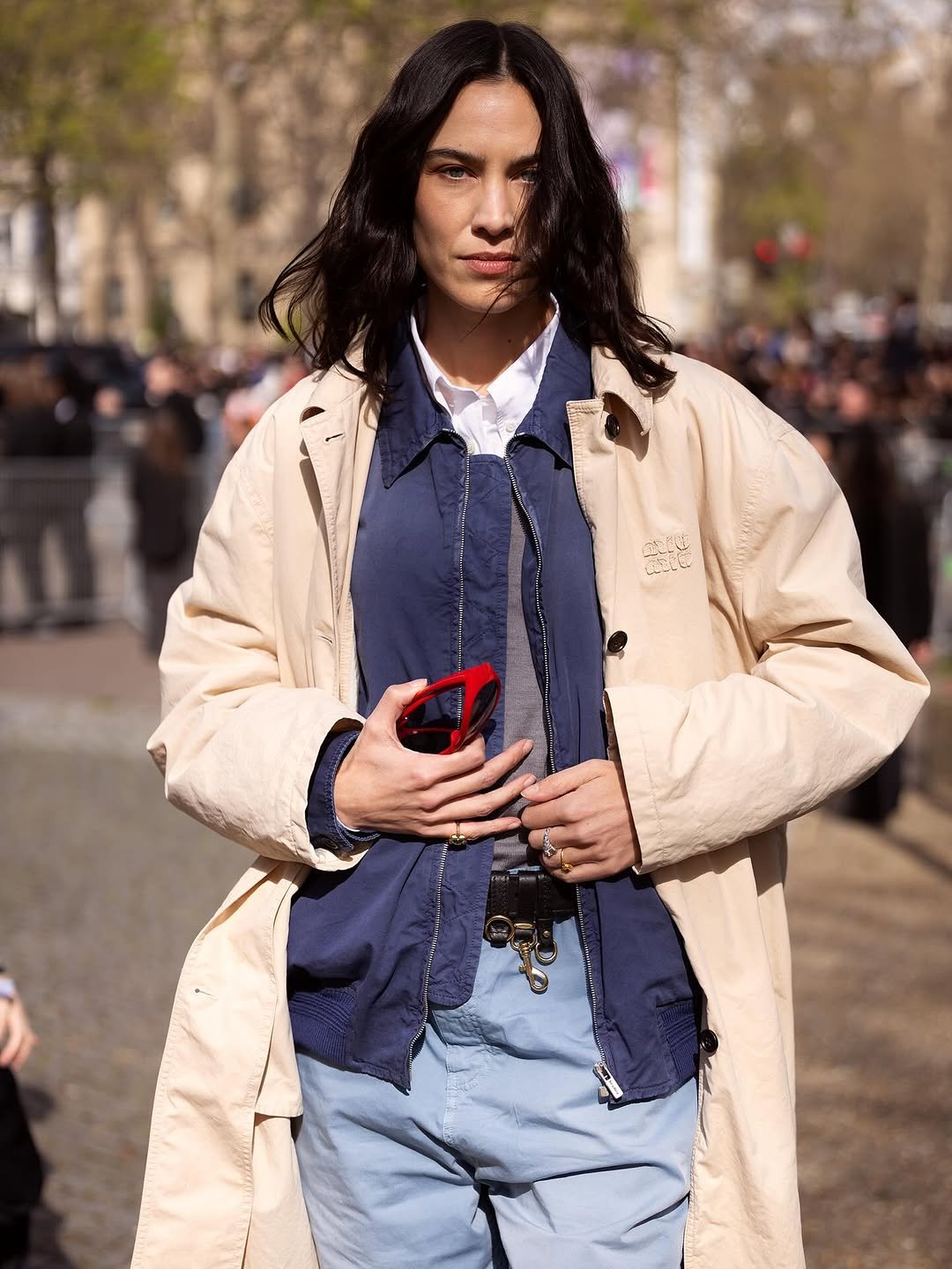 A cream oversized coat is layered over a navy zip jacket, white shirt, grey top, and pale blue trousers with a black belt and rings.