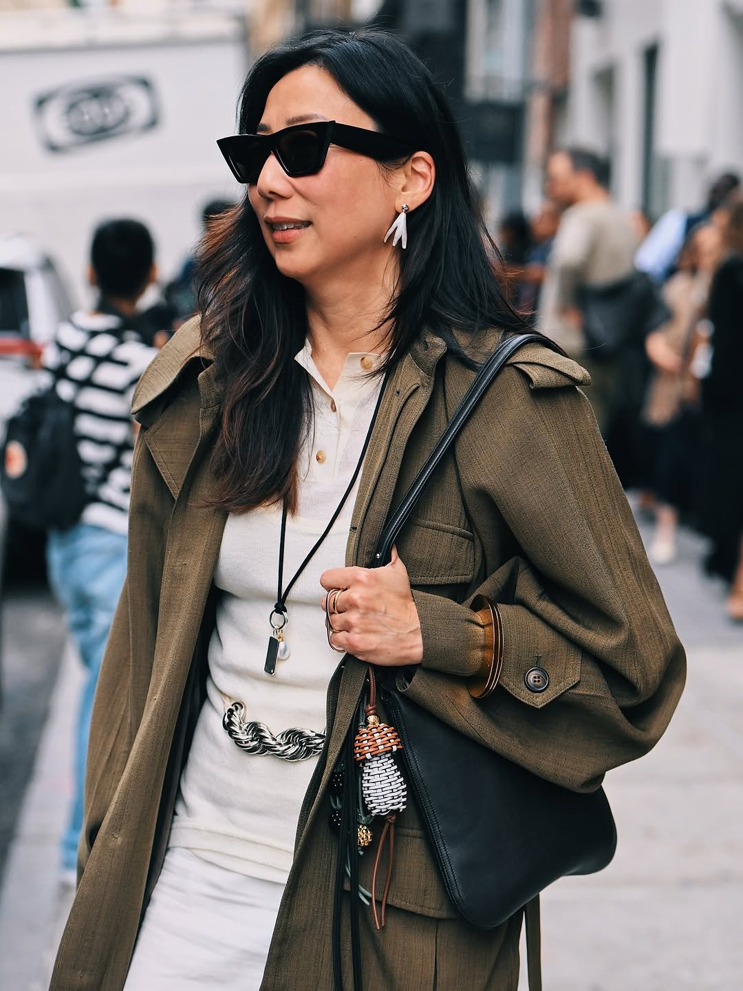 An olive trench-style coat is layered over a cream button knit top with white bottoms, a chain belt, black shoulder bag, sunglasses, and jewelry.