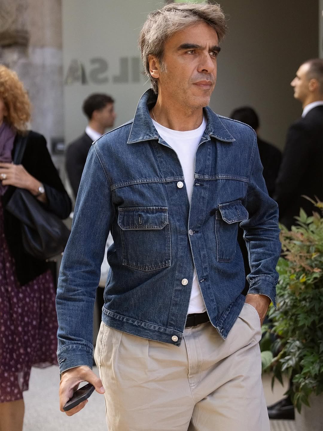 A denim jacket over a white crew-neck T-shirt is styled with beige pleated trousers and a black belt.