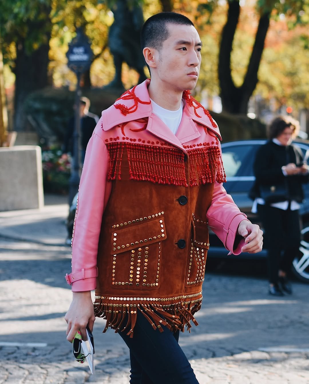 A pink and brown embellished jacket with red beading, gold studs, fringe, a white crewneck layer, and slim dark trousers.