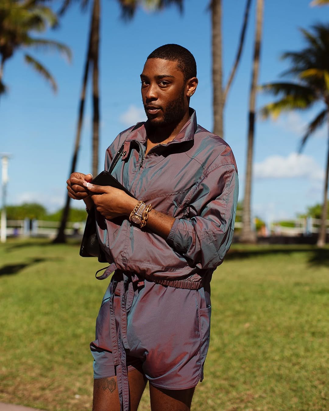 A relaxed iridescent windbreaker romper is styled with gold jewelry and a black shoulder bag.