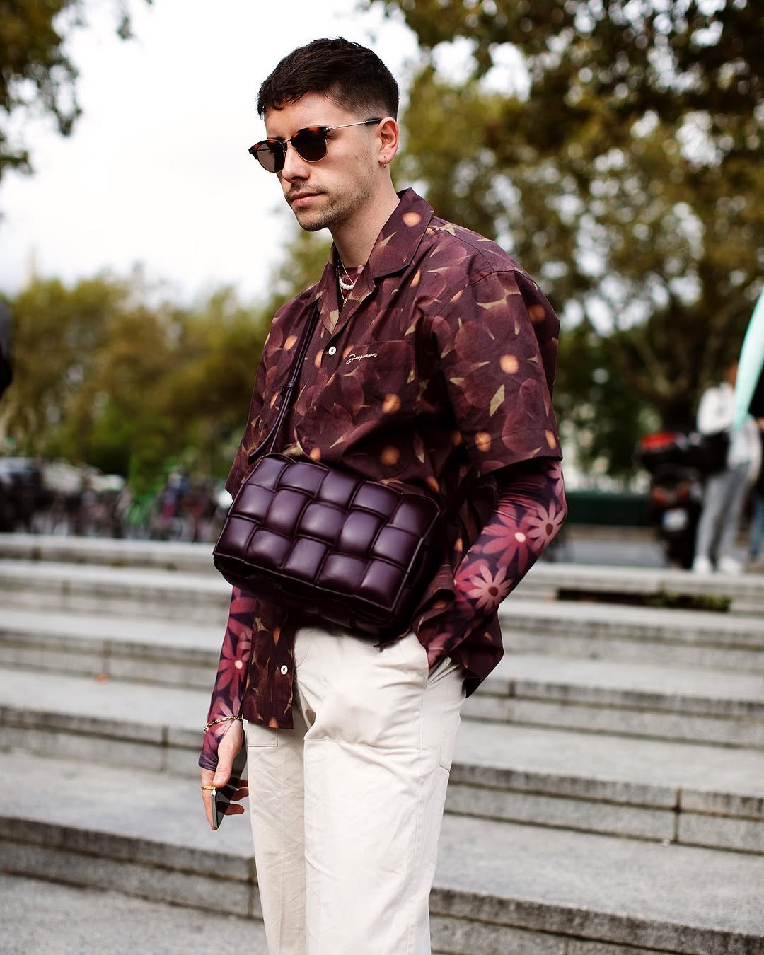 A burgundy floral layered shirt look with cream trousers, dark sunglasses, jewelry, and a padded crossbody bag.