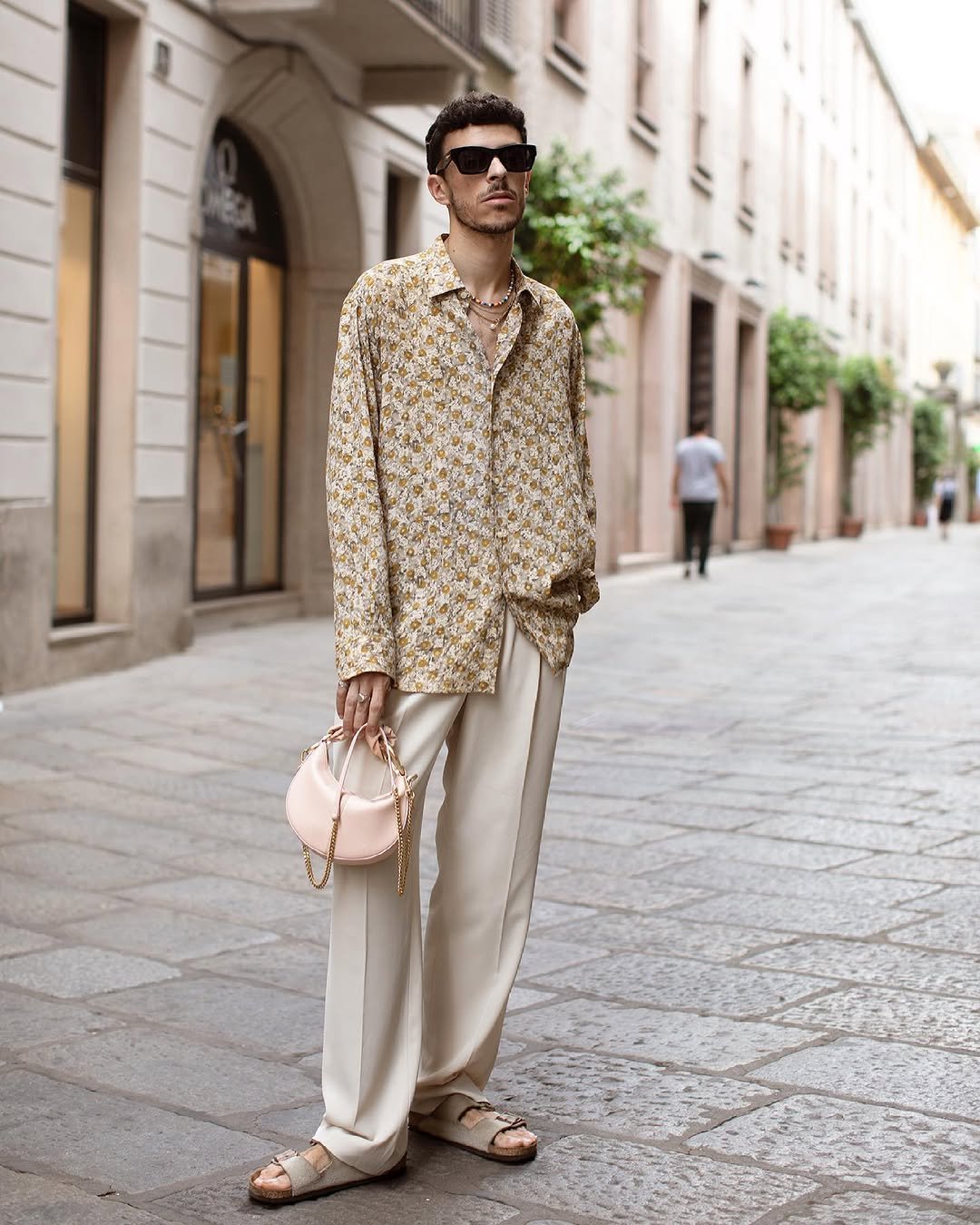 A loose floral shirt is styled open at the neck with wide cream trousers, beige sandals, black sunglasses, layered necklaces, and a pale pink chain bag.