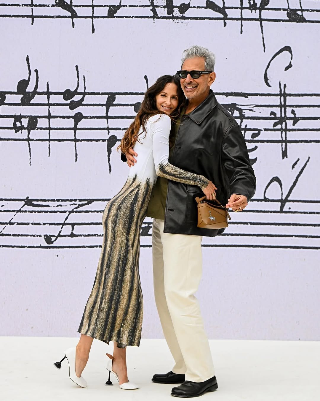 A woman in a white-and-black patterned midi dress and white heels embraces a man in a black leather jacket, olive top, cream trousers, black loafers, and sunglasses.