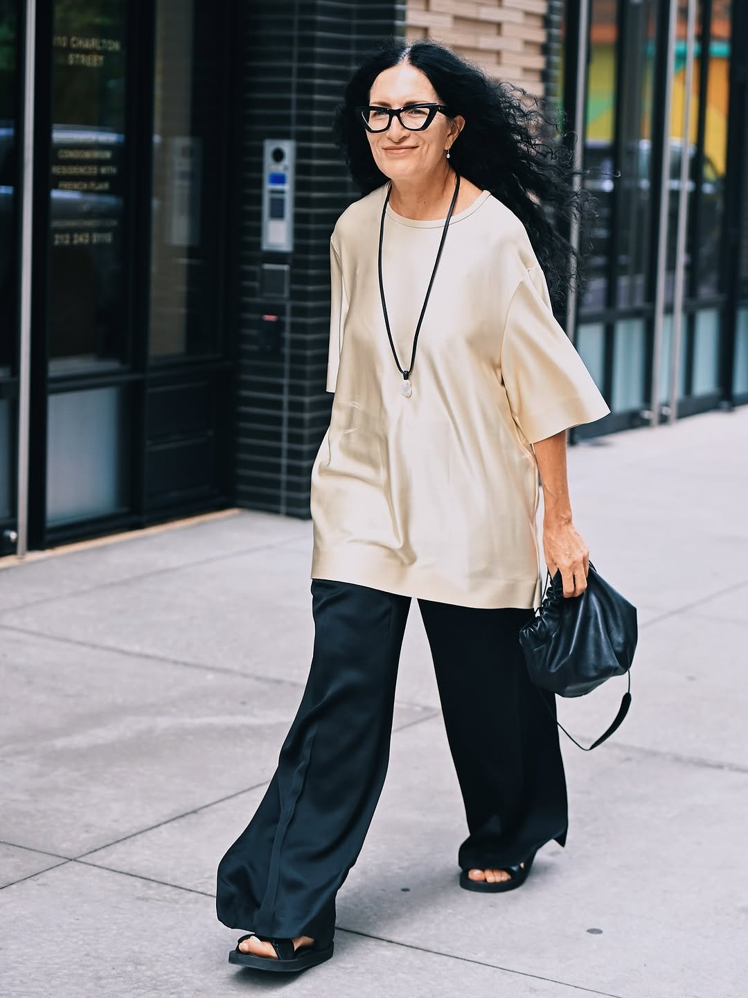 An oversized cream tunic top is styled with wide black trousers, black sandals, a black pouch bag, and a long pendant necklace.