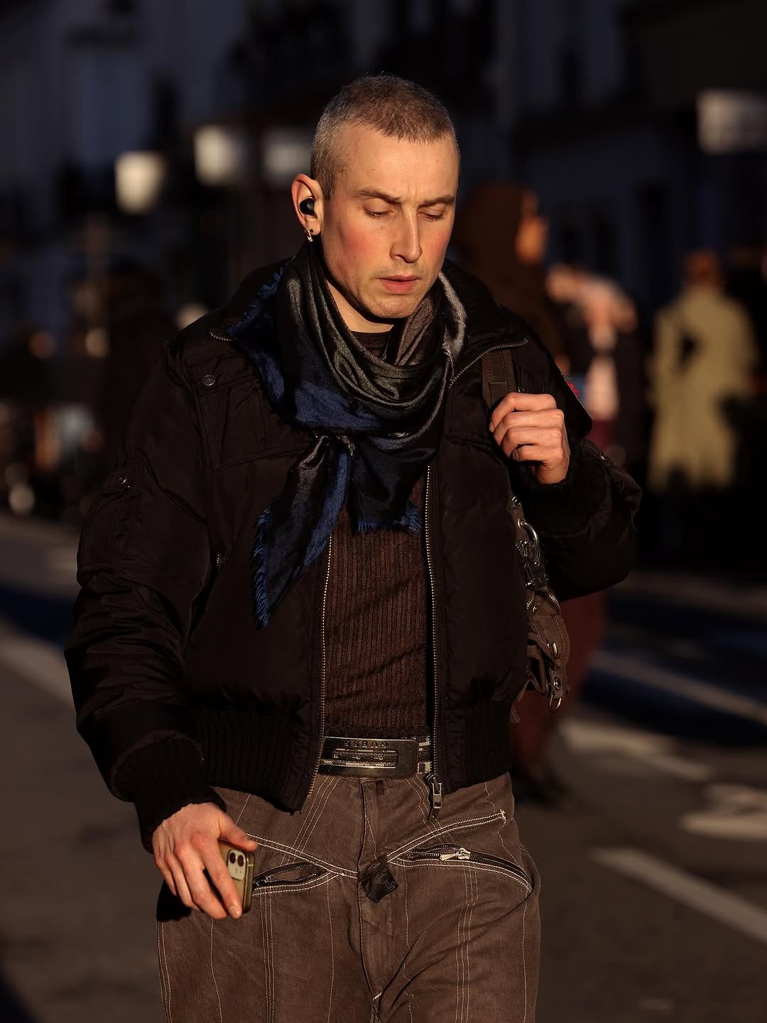 A dark cropped bomber jacket is layered over a ribbed brown knit, a satin scarf, and brown utility trousers with contrast stitching.
