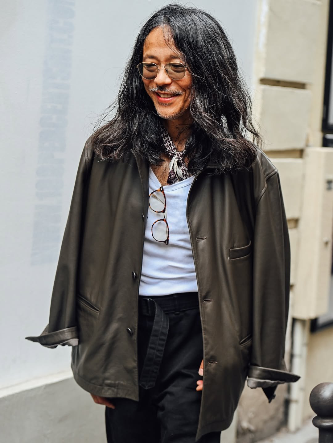 An oversized dark leather jacket is worn over a white ribbed tank, black belted trousers, and a patterned neck scarf.