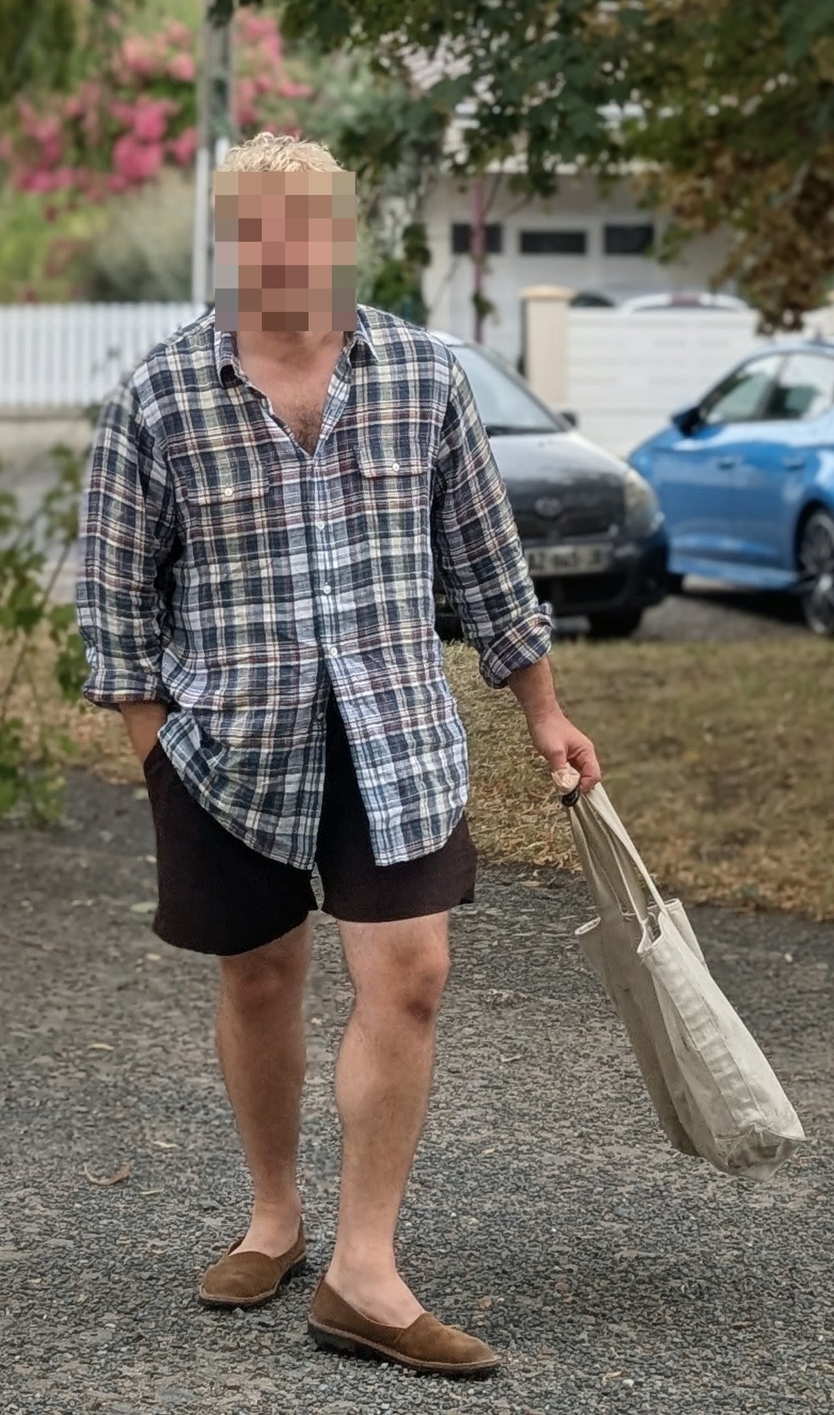 A relaxed plaid button-up shirt is worn open at the neck with black shorts, brown slip-on shoes, and a light canvas tote.