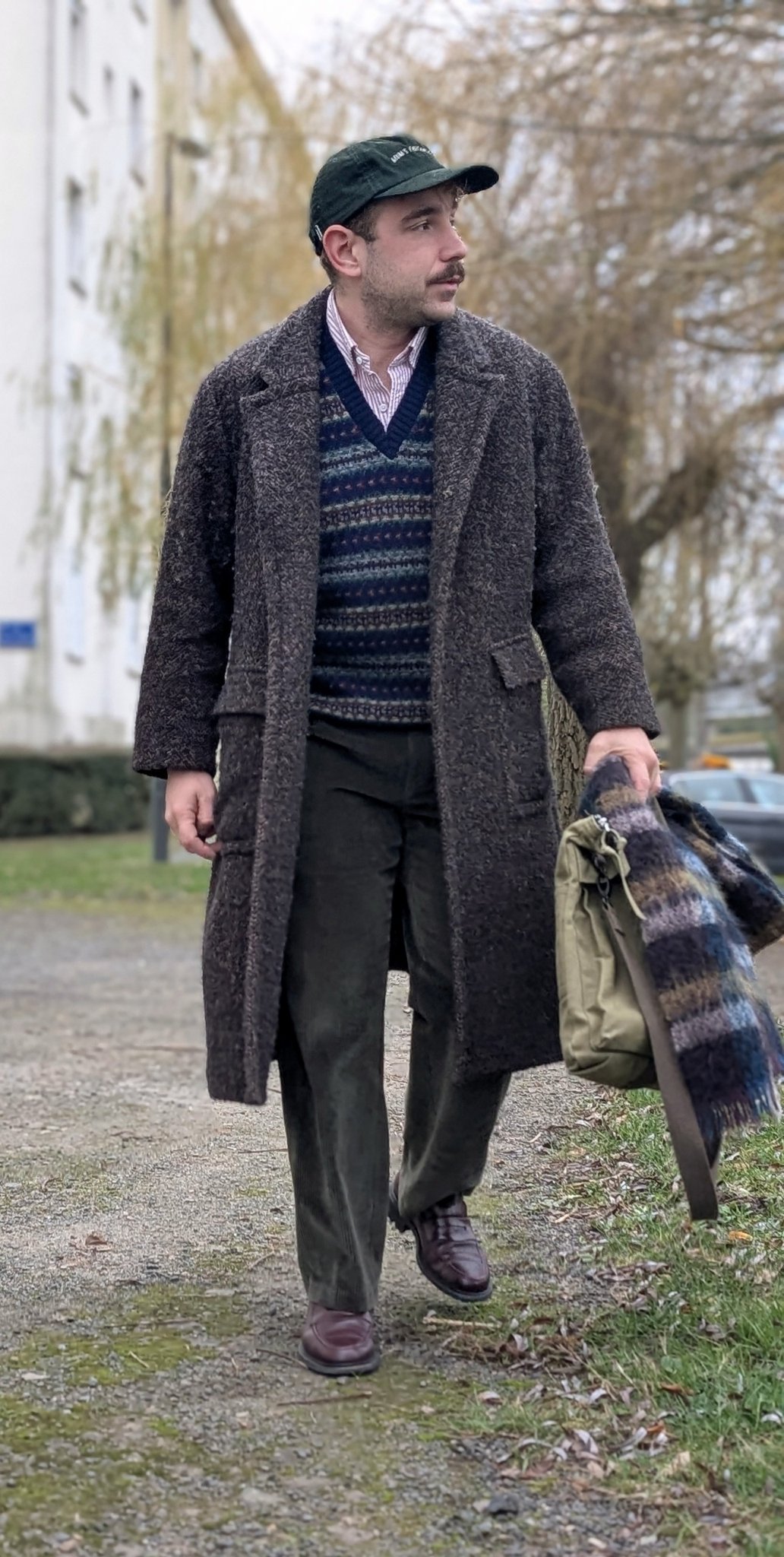 A textured dark overcoat is layered over a patterned V-neck knit, striped shirt, relaxed trousers, brown leather shoes, and a black cap.