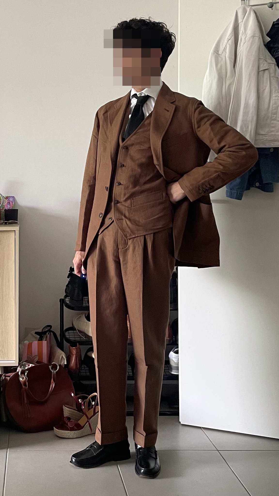 A brown three-piece suit is styled with a white shirt, black tie, and black leather loafers.