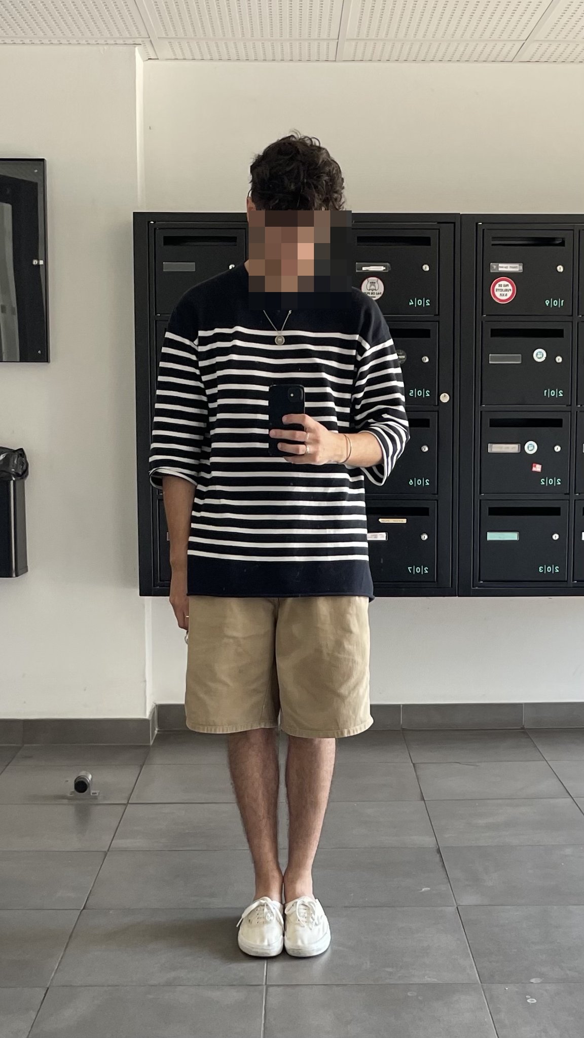 A relaxed black-and-white striped oversized top is styled with tan knee-length shorts, white low-top sneakers, and a small pendant necklace.