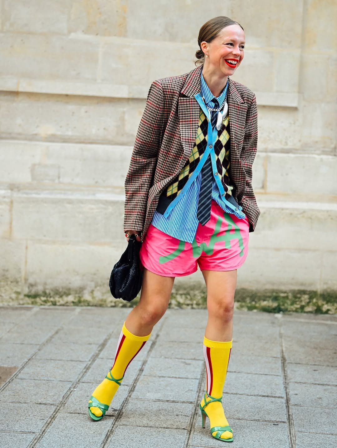 A layered print-heavy blazer, striped shirt, argyle cardigan, tie, pink graphic shorts, yellow knee socks, and green heeled sandals create a bold preppy-street look.