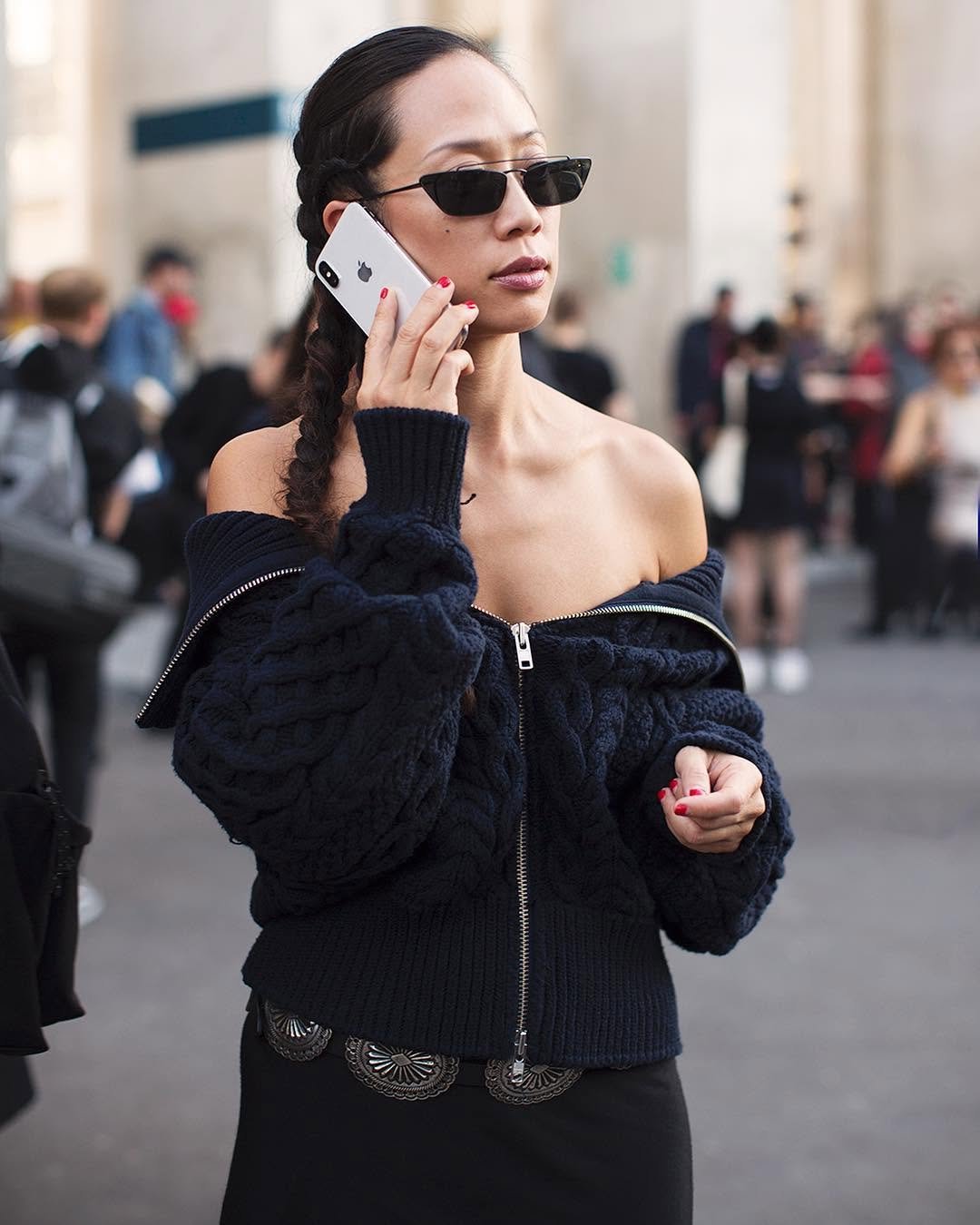 A navy chunky cable-knit zip cardigan is worn off the shoulders with black bottoms and narrow cat-eye sunglasses.