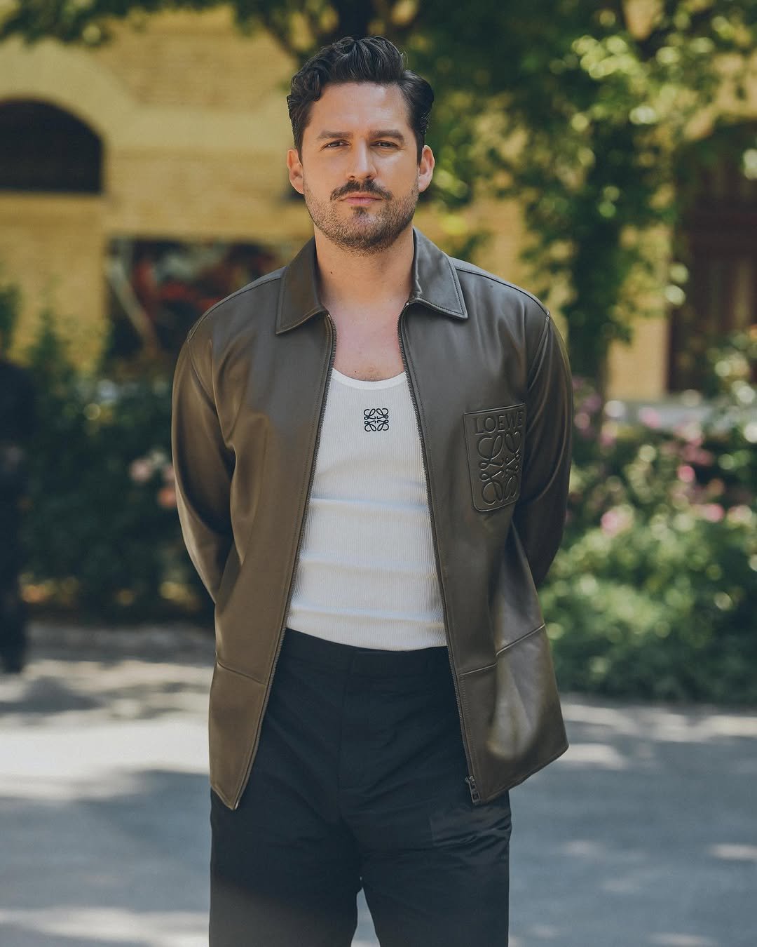 A dark olive leather zip jacket is worn open over a white ribbed tank with black trousers.