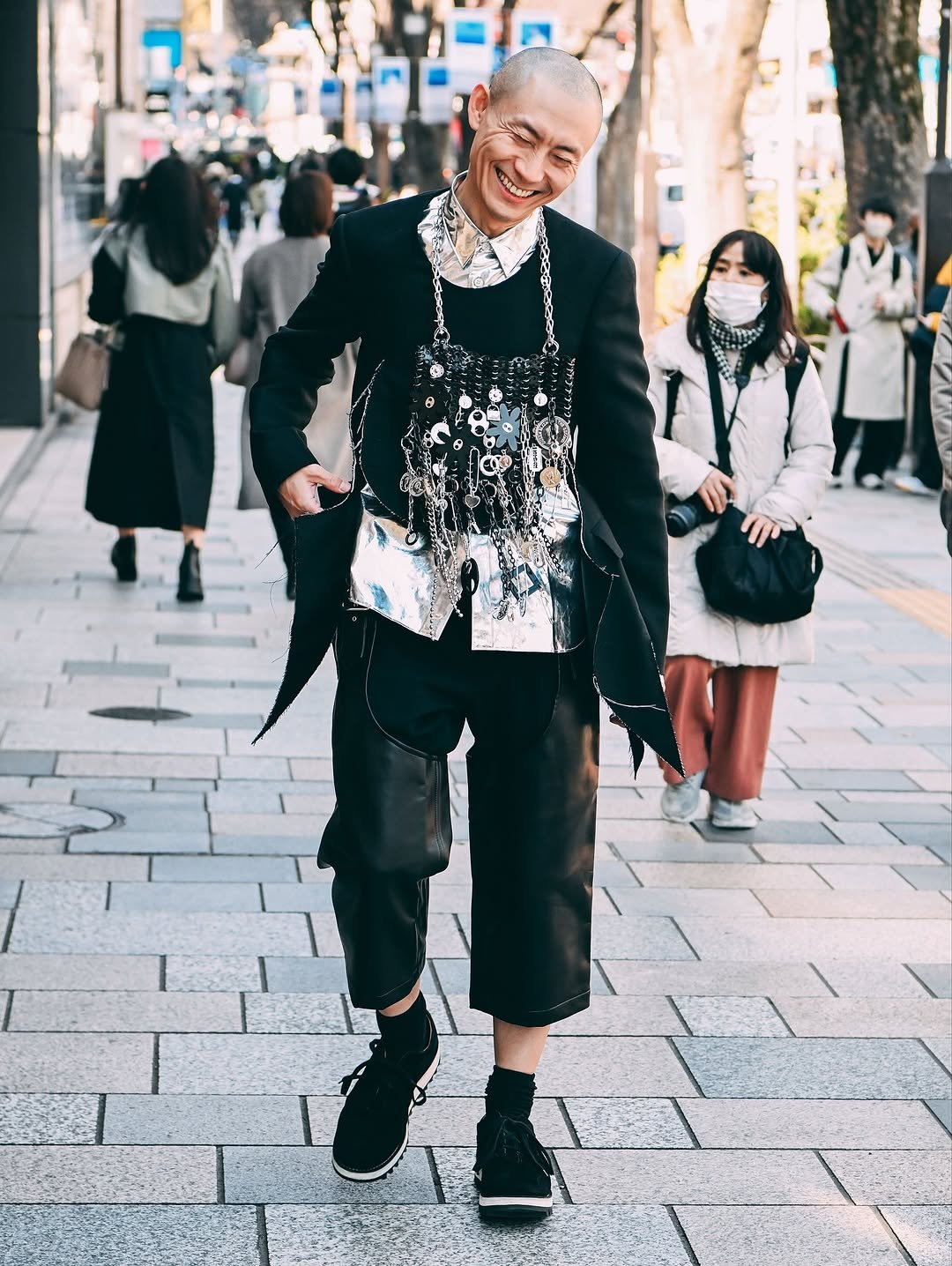 A black tailored jacket and cropped wide shorts are layered with a reflective silver shirt and a dense chain-and-charm chest piece.