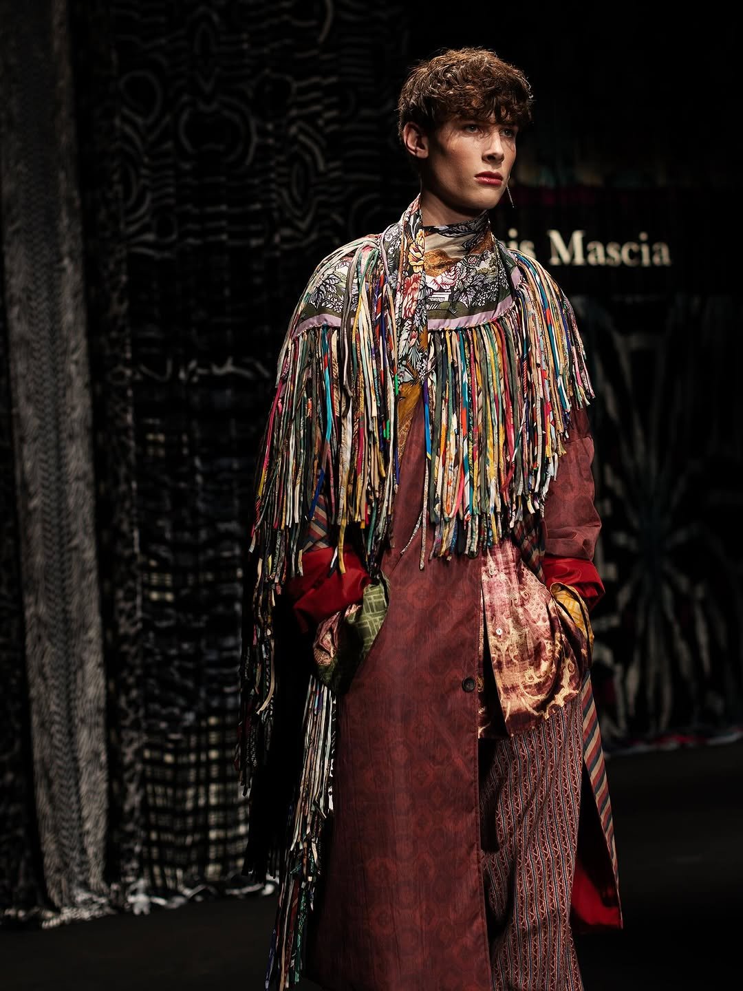 A layered runway look mixes a rust patterned long coat with multicolor fringe, printed shirting, scarfing, and patterned trousers.