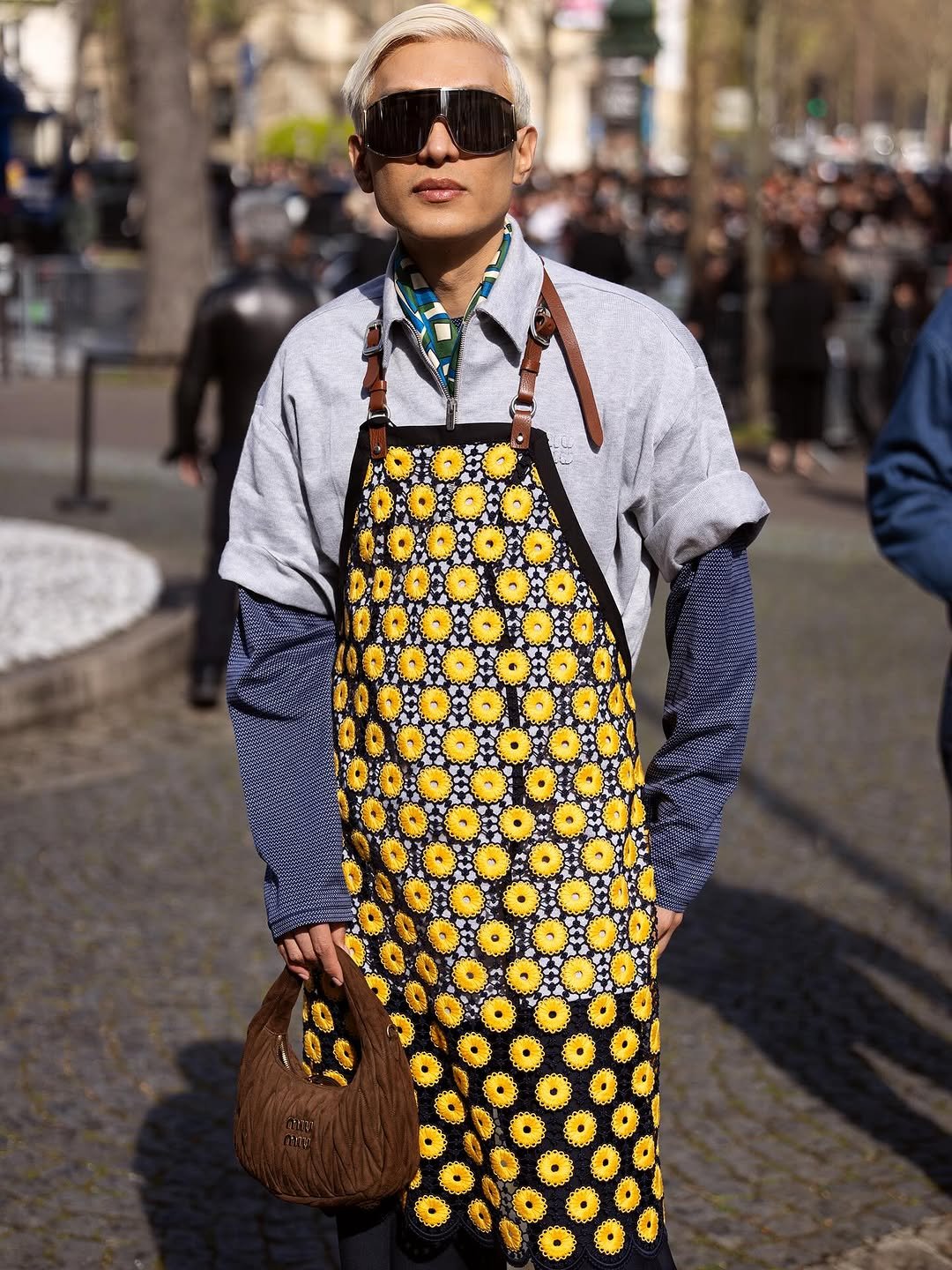 A sunflower-patterned apron dress is layered over a pale grey short-sleeve zip shirt, blue long sleeves, oversized visor sunglasses, and a brown quilted handbag.