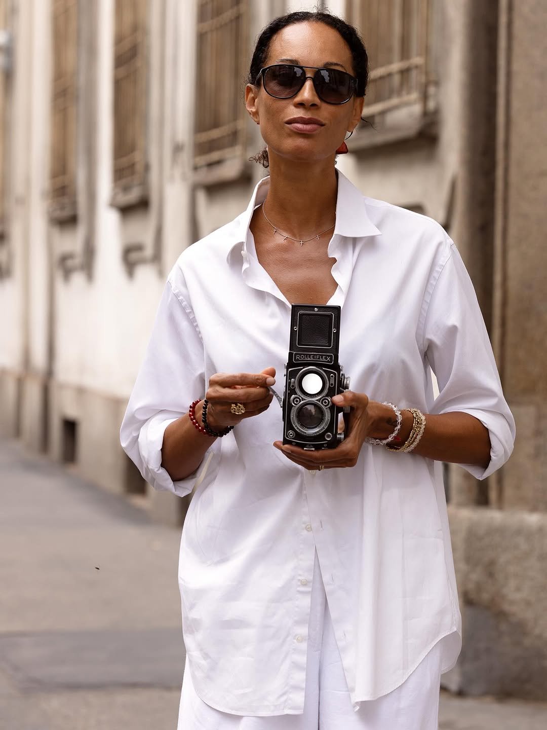 A crisp white oversized button-up shirt is styled with dark sunglasses, layered bracelets, a delicate necklace, and white lower layers.