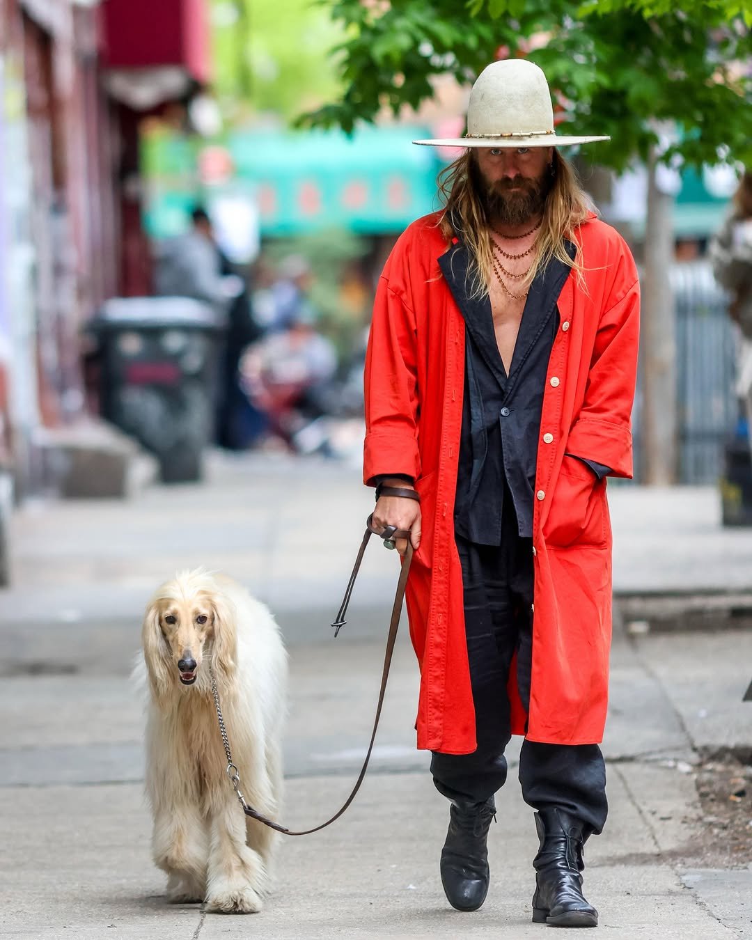 A bright red long coat is layered over a black open shirt, black trousers, tall black boots, and a wide-brim cream hat.