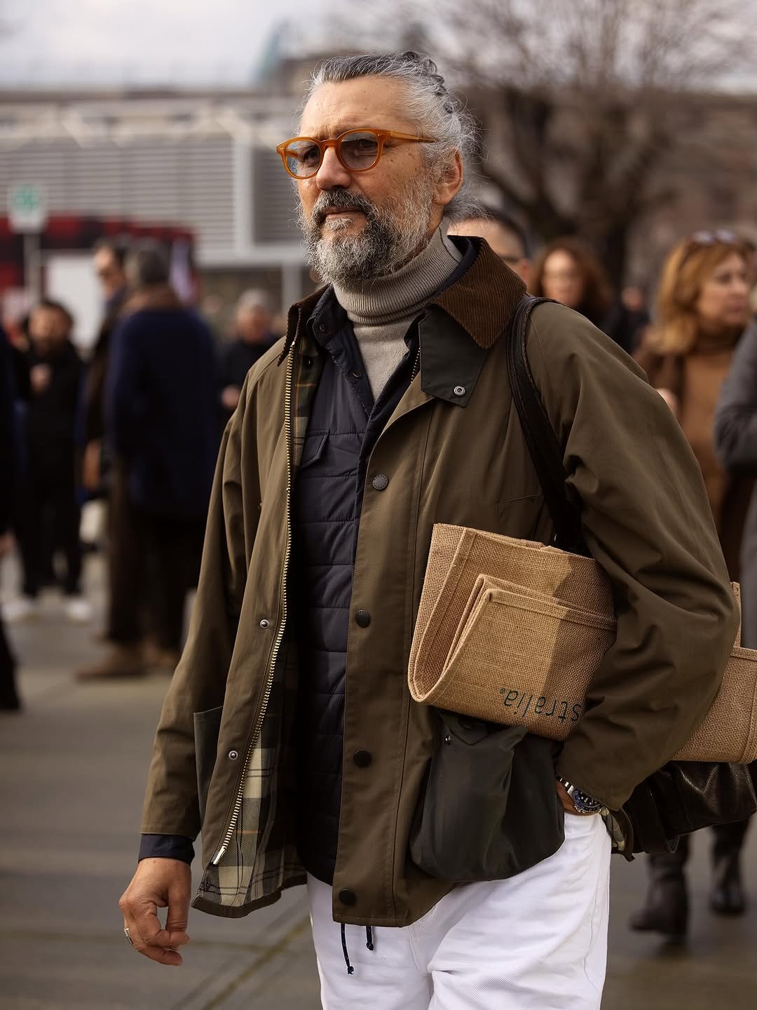 A layered olive field jacket look with a grey roll-neck, dark quilted vest, white trousers, amber sunglasses, and a tan woven tote.