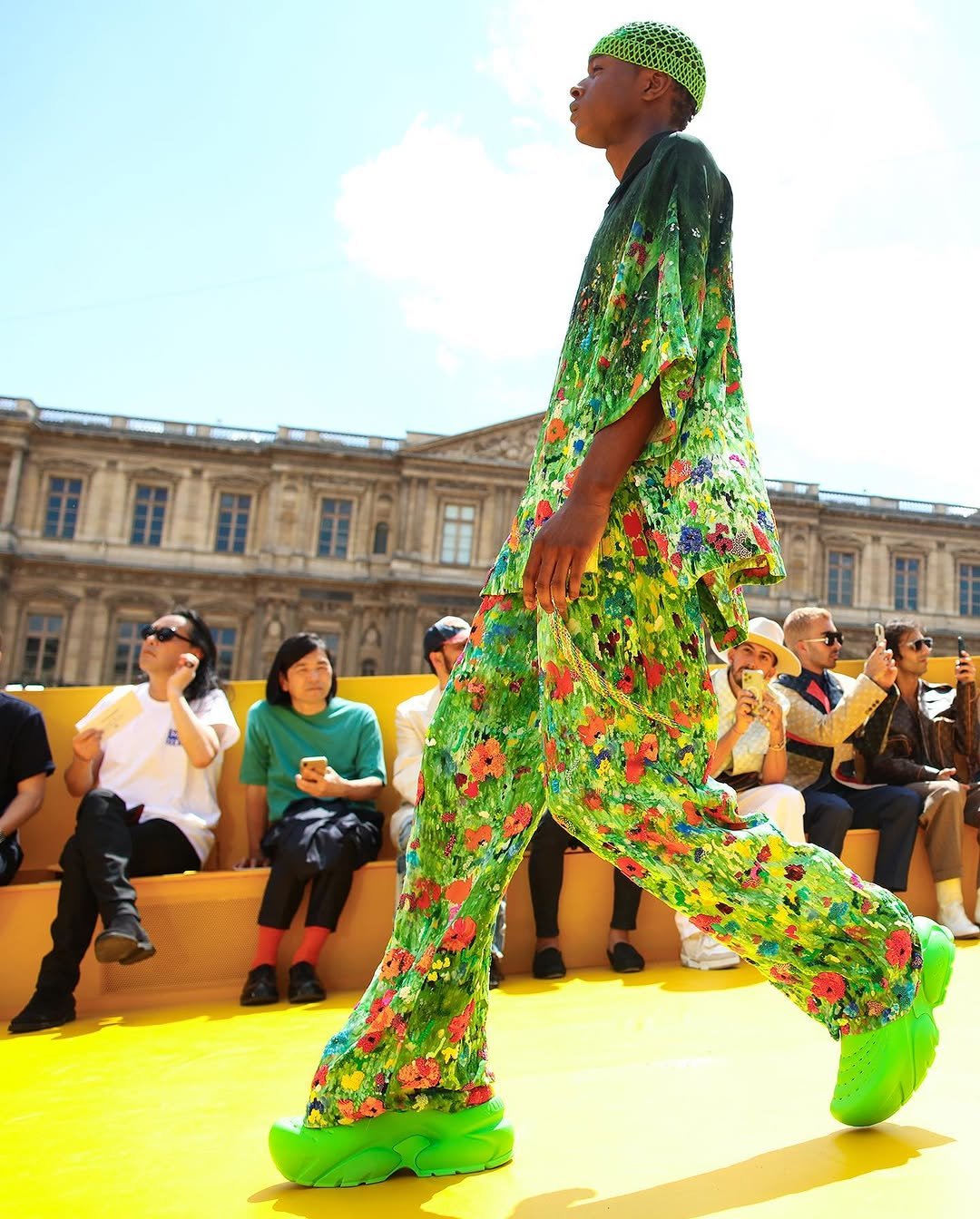 A model wears a vivid green floral oversized top and matching flared trousers with neon green chunky shoes and a mesh cap.