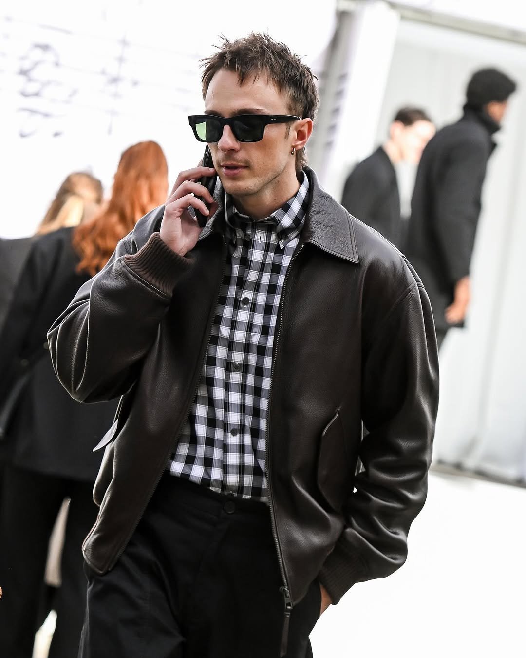 A dark leather blouson jacket is worn over a black-and-white checked shirt with black trousers and rectangular sunglasses.