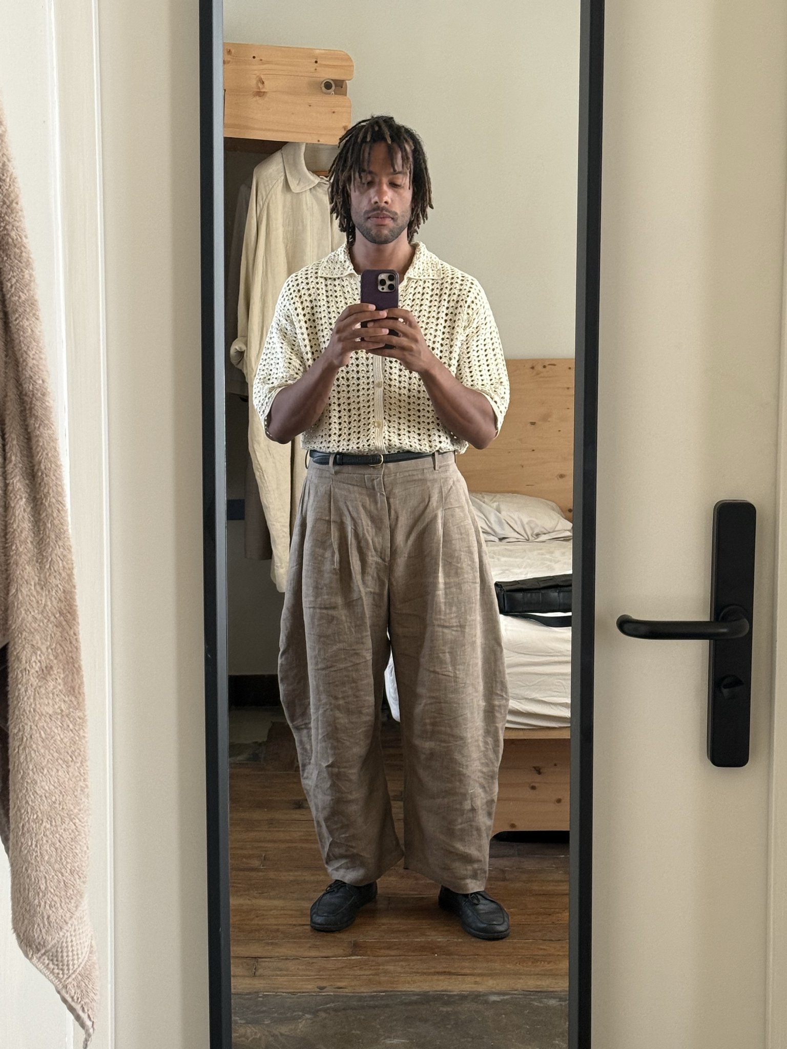 Cream open-knit collared shirt tucked into voluminous beige pleated trousers with a black belt and black shoes.