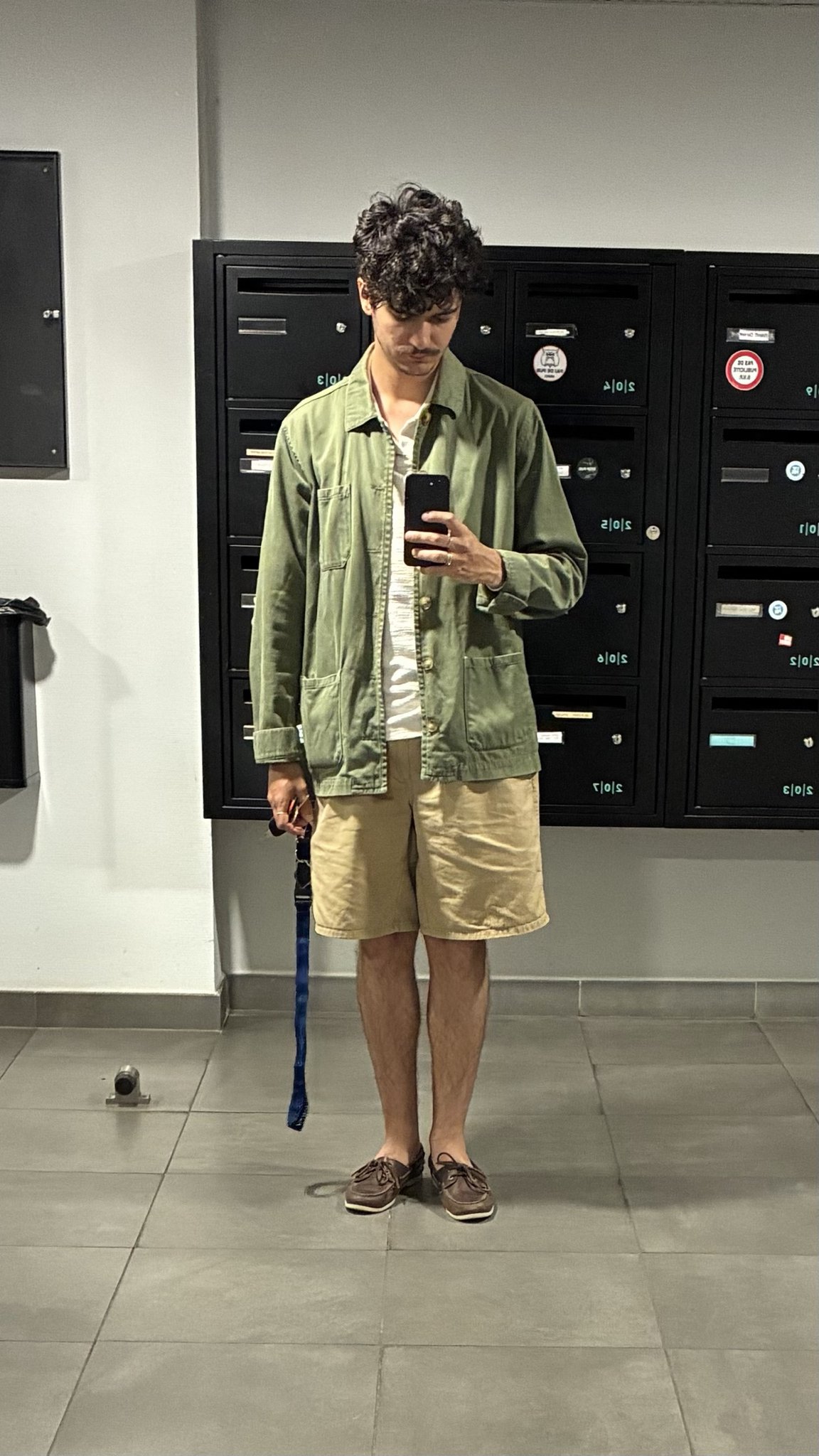 A relaxed olive chore jacket is layered over a white T-shirt with tan shorts and brown boat shoes.